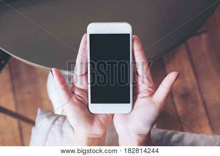 Mockup image of hands holding white mobile phone with blank black screen on pillow with wooden floor background