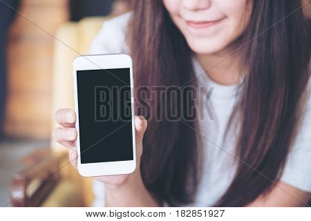Mockup image of a beautiful woman holding and showing white mobile phone with blank black screen with smiley face in vintage wooden cafe