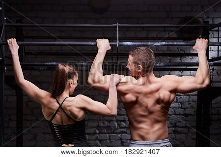 Rear view photo of muscular man and woman doing exercises on horizontal bar against brick wall at the cross fit gym. Sporty couple in the sportwear. Power and energy. Healthy lifestyle. Concept of the cross fit activity in the couple.