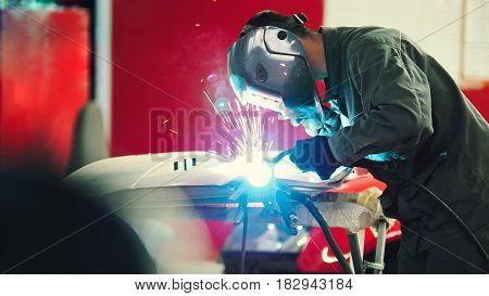 Welding industrial: worker in helmet repair detail in car auto service - blue sparklers, telephoto