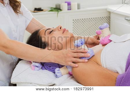 Herbal ball massage in ayurveda spa. Female massagist with young woman in wellness center. Healthcare therapy to beautiful indian girl in beauty parlor, side view of face with eyes closed