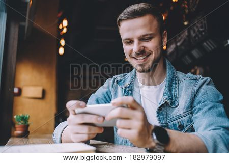 Young handsome smiling man looking at phone sitting at the restaurant near the window. Attractive guy relaxing having fun. Close-up