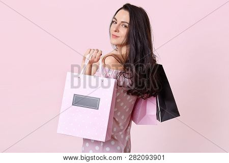 Good Looking Brunette Woman Stands Sideways, Holds Shopping Bags, Looks Directly At Camera, Returns 