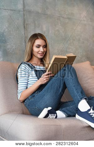 Young Smiling Girl Sitting On Pink Sofa And Reading Book