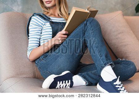 Cropped View Of Young Woman Sitting On Pink Sofa And Holding Book