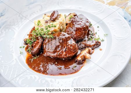 Traditional German braised pork cheeks in brown red wine sauce with mushroom and mashed potatoes as closeup on a classic design white plate 