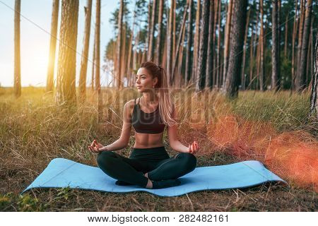 Beautiful And Young Girl Sitting On The Rug In The Lotus Position. Practicing Yoga In Asana Position