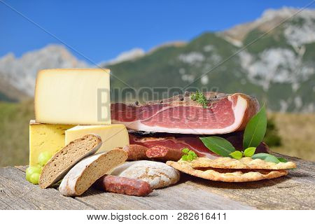 South Tyrolean Specialties Like Bacon, Sausages And Cheese Lying On A Rustic Table In Front Of Mount