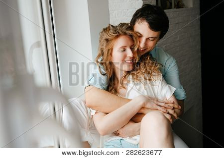 Happy Girl And Man Hugging Near Window In Home. White And Blue Clothes. Valentine Day.