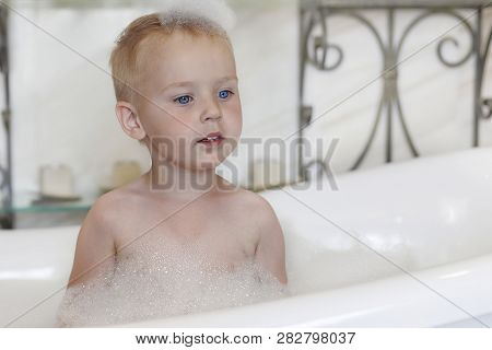 Cute Caucasian Baby Boy With Bright Blue Eyes And Fair Hair In The Bathtube, With Foam Bubbles O The