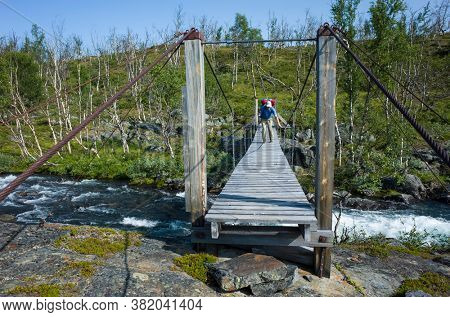 Hiking in Sweden in summer. Man crossing river on wooden suspension bridge, Trekking alone in Abisko National Park in Lapland. Arctic nature of Scandinavia in warm sunny day