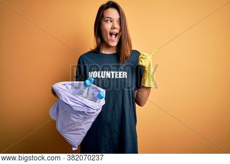 Beautiful volunteer girl caring environment doing volunteering holding bag with rubish bottles pointing and showing with thumb up to the side with happy face smiling