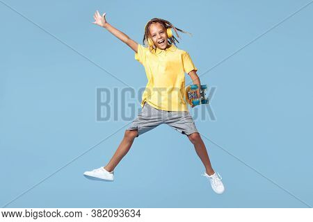 Happy Little Boy With African Dreads With Headphone And Skateboard Jumping Over Blue Background.