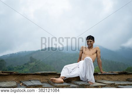 Young Shredded Man Sitting Wearing A Dhoti And Glasses With Blurred Mountains In The Background.