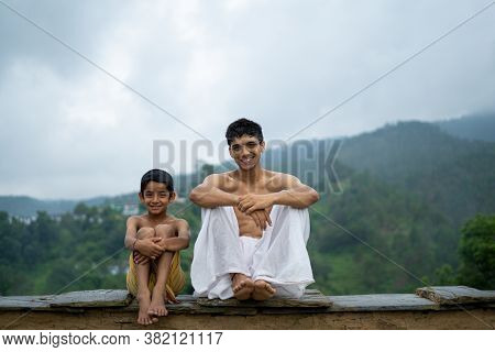 A Young Man Sitting On The Roof With His Younger Brother,both Wearing Dhoti, Smiling Into The Camera