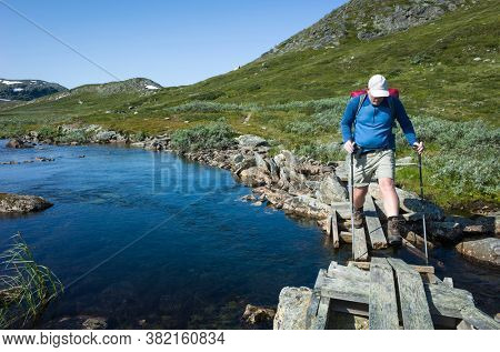 Hiking in Swedish Lapland. Man crossing river on small wooden broken bridge, Trekking alone in north Sweden. Arctic nature of Scandinavia in warm summer sunny day with blue sky