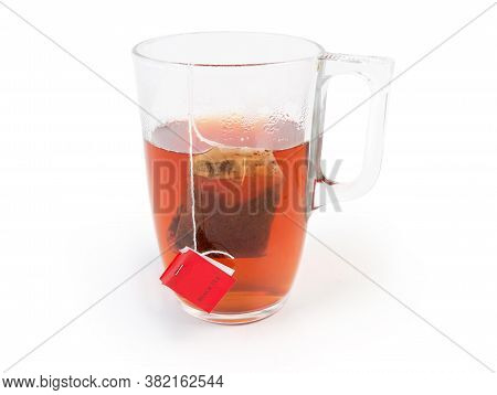 Bag Of Black Tea With Red Label In The Glass Cup With Hot Freshly Brewed Tea On A White Background