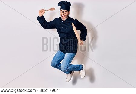 Middle age handsome hispanic man wearing glasses and cook uniform smiling happy. Jumping with smile on face holding kneader and spoon over isolated white background