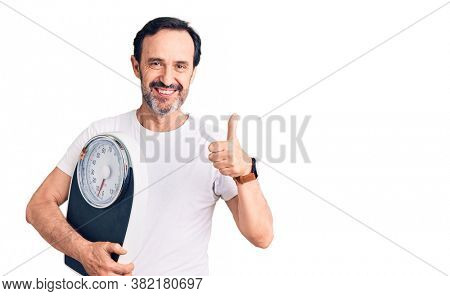 Middle age handsome man holding weighing machine smiling happy and positive, thumb up doing excellent and approval sign 