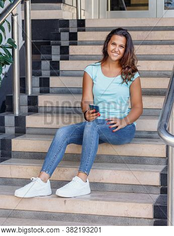 Young Women With Mobile Phone Sits On The Steps And Smiling