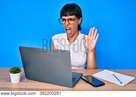 Beautiful brunettte woman working at the office doing video call angry and mad screaming frustrated and furious, shouting with anger. rage and aggressive concept. 