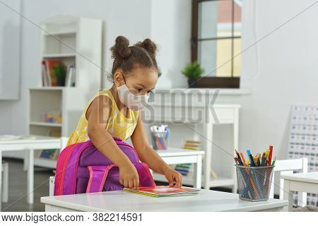 An African-american Girl With Medical Mask On Her Face Puts A Copybook On A Schooldesk. A Child In A