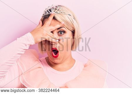 Beautiful blonde plus size woman wearing pincess tiara with diamonds over pink background peeking in shock covering face and eyes with hand, looking through fingers afraid