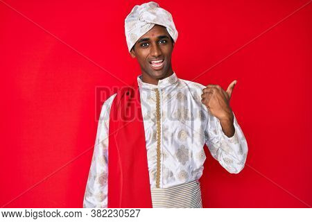 Handsome indian man wearing tradition sherwani clothes smiling with happy face looking and pointing to the side with thumb up. 
