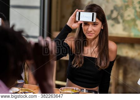 A Beautiful Girl With Her Smartphone On Her Forehead Sitting At The Table With Friends And Trying To