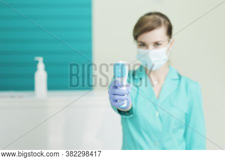 Female Doctor Or Nurse In Protective Mask At Hospital Holds Pyrometer In Hand. Safety Measures Again