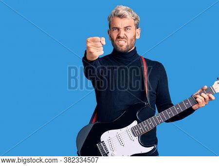 Young handsome blond man playing electric guitar annoyed and frustrated shouting with anger, yelling crazy with anger and hand raised 