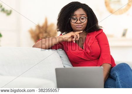 Portrait Of Confused Skeptic Black Curly Woman In Eyeglasses Looking At Her Laptop Sitting On Couch 