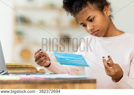 School During Pandemic. Black Teen Girl Holding Protective Mask Sitting At Desk Indoors. Selective F