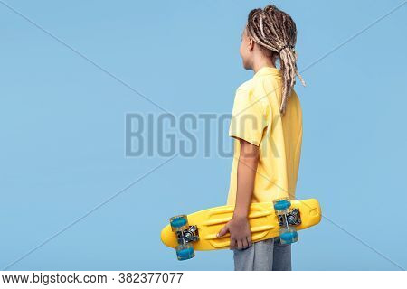 Stylish Little Boy With African Dreads With Skateboard On The Blue Background. Back View.