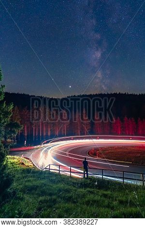 Milkyway, Long Exposure Of A Man Standing At A Railing In Front Of A Curvy Street, While Cars Are Pa