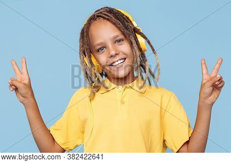 Cheerful Little Boy With African Dreads Listening Music In Headphones Showing Peace Gesture Over Blu