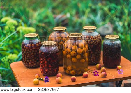 Jars With Compote On Table. Sealed Glass Jars With Kompot Placed On Table Near Flowers And Fruits On