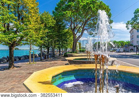 Fountains And Green Trees On Promenade Embankment Of Garda Lake In Historical Centre Of Desenzano De