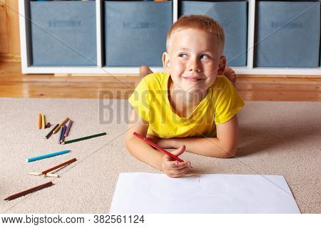 A Blond Boy In A Yellow T-shirt Lies On The Floor With A Drawing Sheet And Pencils And Smiles Though