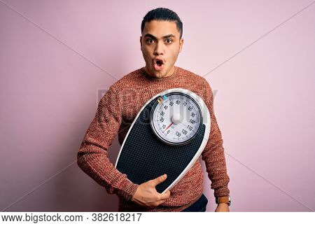 Young brazilian man doing diet to lose weigth holding scale over isolated pink background scared in shock with a surprise face, afraid and excited with fear expression