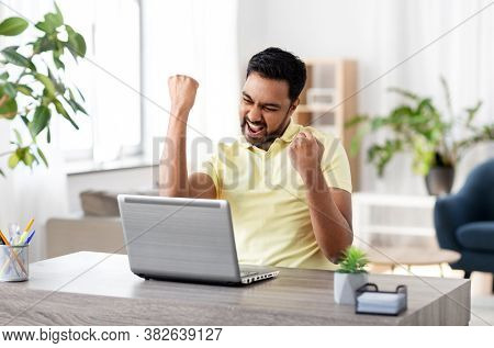 technology, remote job and lifestyle concept - happy smiling indian man with laptop computer working and celebrating success at home office