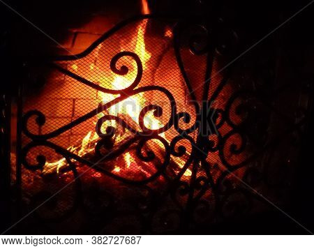 Fire In The Fireplace Behind A Beautiful Wrought Iron Grate With Curls. A Fire Is Burning In The Fir
