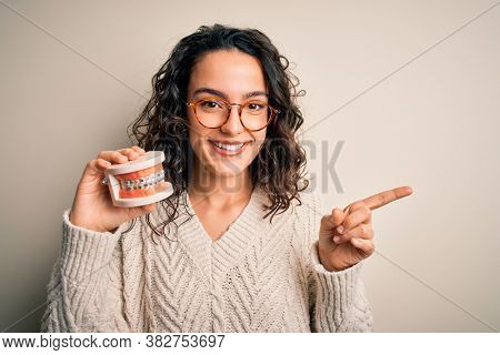 Young beautiful woman with curly hair holding plastic teeth with dental baces very happy pointing with hand and finger to the side