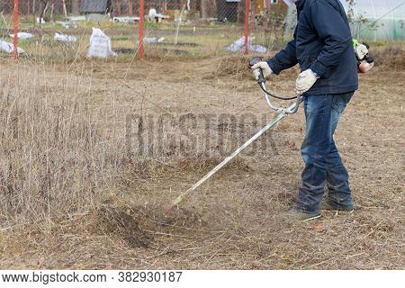 Man Wearing A Blue Coat And Jeans Is Trimming The Grass. Mowing A Grass. Rural Activities. Selective
