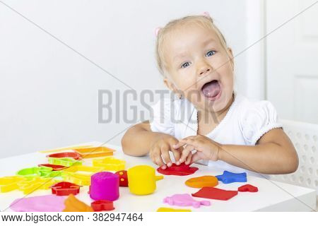 Toddler Sculpts From Colored Plasticine On A White Table. The Hand Of A Small Child Squeezes Pieces 