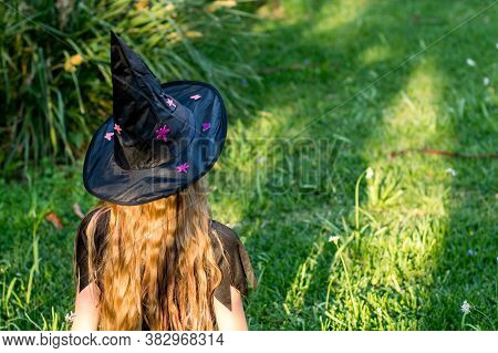 Causcasian Blond Girl With Long Hair In Halloween Witch Costume Sitting On A Green Grass