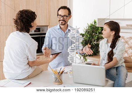 Pleasant talk. Charming little girl sitting on the kitchen counter as well as her brother holding a tablet and talking with the father making explanatory gesture