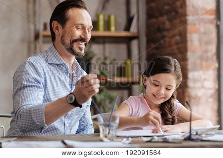 Inspiring co-creation. Joyful young father holding a brush, being ready to apply some strokes on the picture thus helping his cute little daughter to finish up a picture