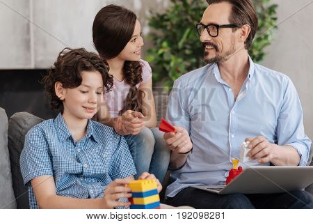 Here you go. Pleasant young father handing a piece of an erector set to his charming smiling daughter while his handsome son assembling an erector set