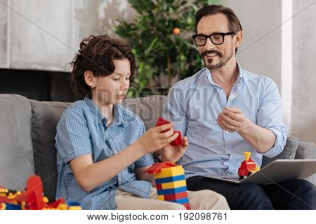 Maximum concentration. Cute serious boy sitting on the sofa and being engaged into assembling erector set into a tower while his father addressing him with a smile